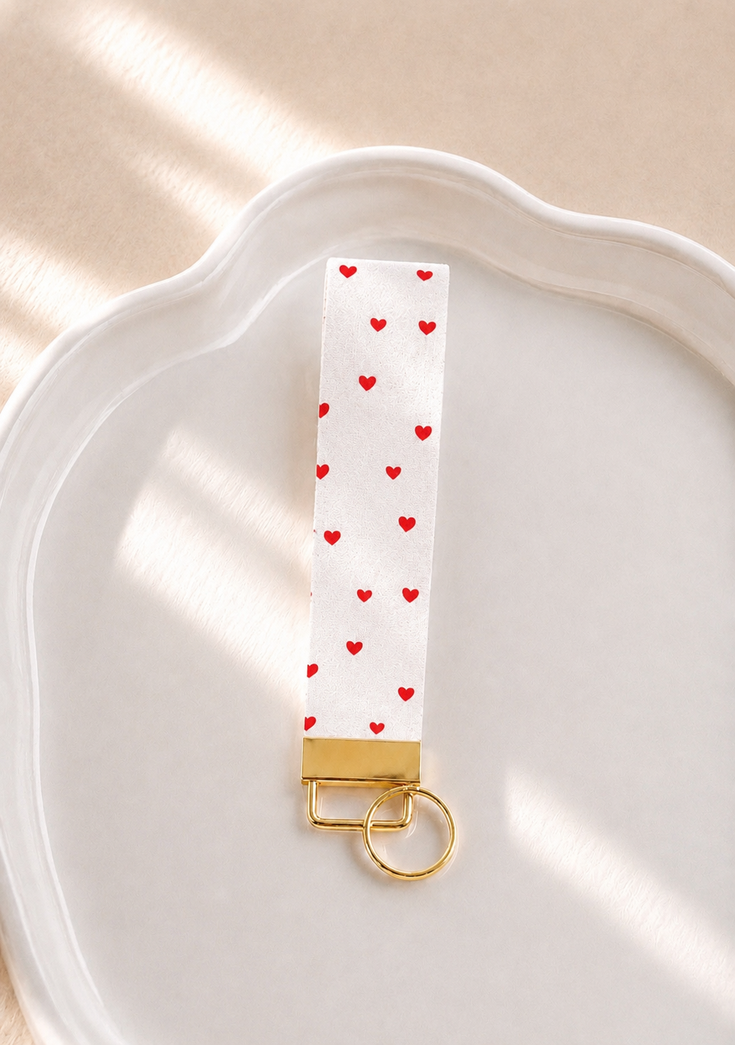 Love You Wristlet keychain with white and red heart print and gold hardware laid flat on a white ceramic dish