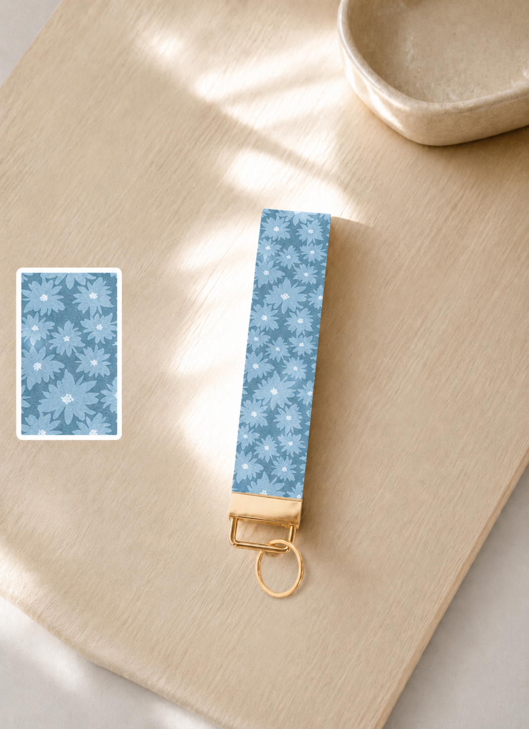 Blue Skies wristlet keychain with blue daisy print and gold hardware, laid flat on wood surface in natural light with print detail inset