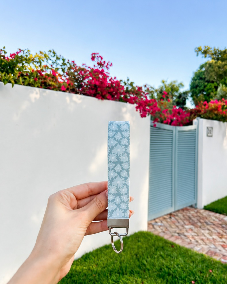 Hand holding Blue Skies wristlet keychain with powder blue daisy print and silver hardware, outdoors with bougainvillea and blue gate in background