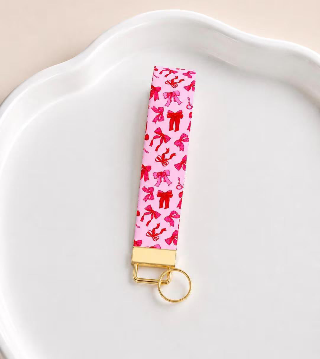 Blush Bows wristlet keychain with light pink base covered in scattered red and hot pink bows, laid on white scalloped tray with gold hardware