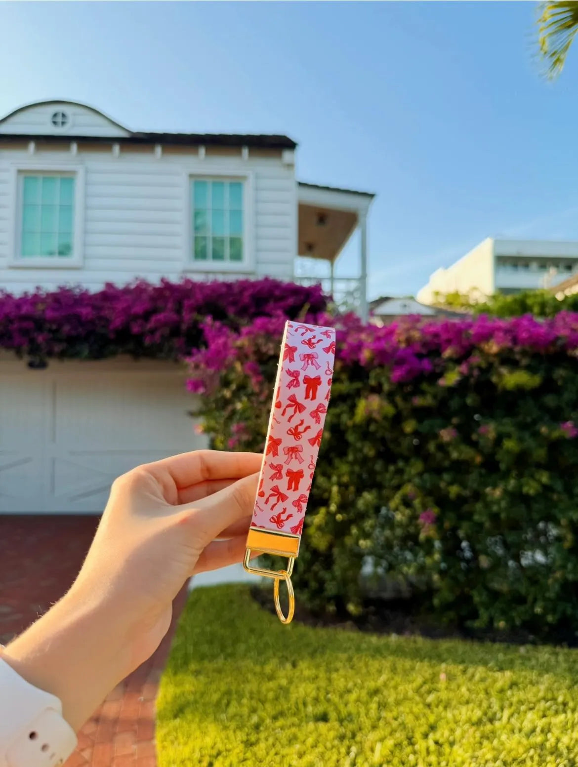 Hand holding Blush Bows wristlet keychain with light pink and red bow print outdoors with pink bougainvillea in background and gold hardware