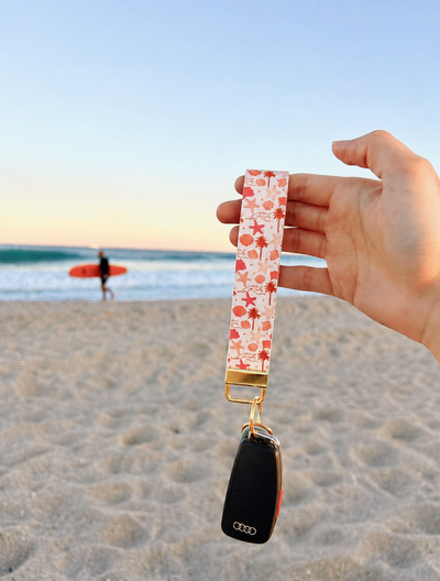 A hand holds a car key fob wrapped in a white fabric wrist strap with a red and orange floral pattern, set against a blurred beach background with ocean waves and a surfer in the distance.