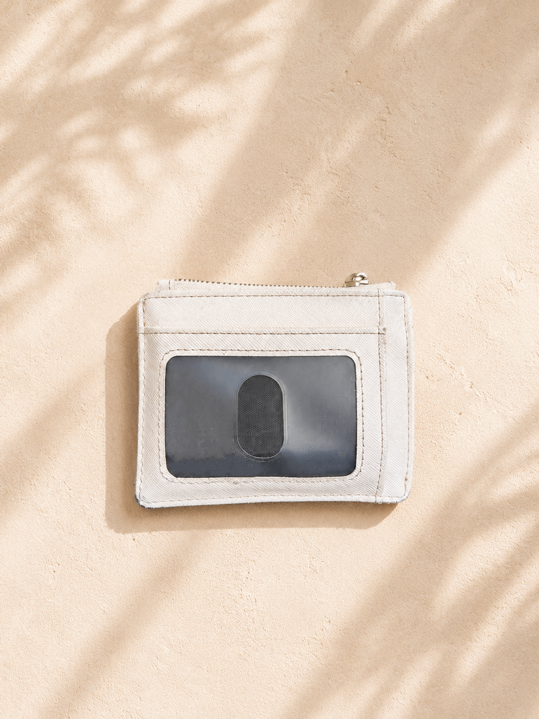 A slim white leather card holder wallet with a clear ID window, shown on a textured beige surface with soft leaf shadows.