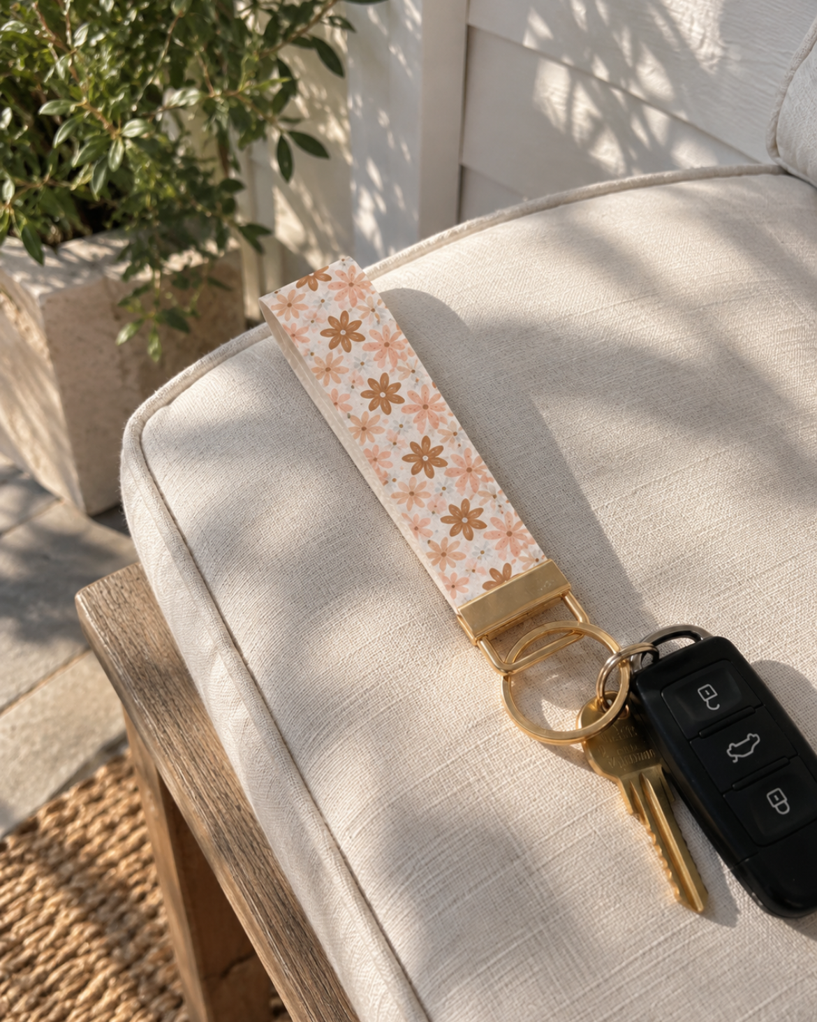 A keychain with a cream and orange floral patterned strap and gold-toned hardware, holding car keys, resting on a light beige outdoor chair cushion.