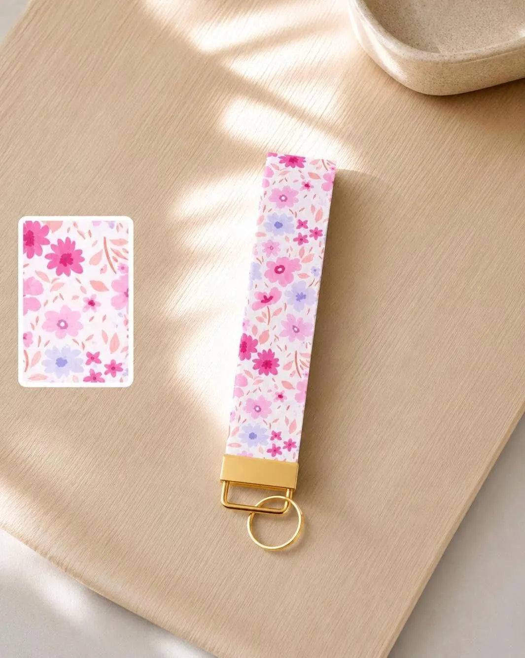 Floral Daydream wristlet keychain with hot pink, blush, lavender, and periwinkle ditsy daisy print on white, laid on wood surface in natural light with print detail inset and gold hardware