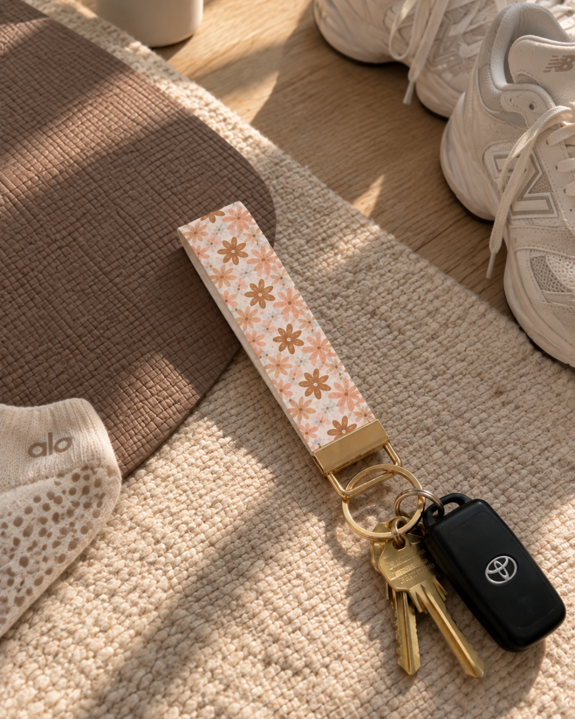 A close-up of a floral-patterned fabric keychain with a gold-tone keyring holding keys, placed on a textured rug near white sneakers in a styled indoor setting.