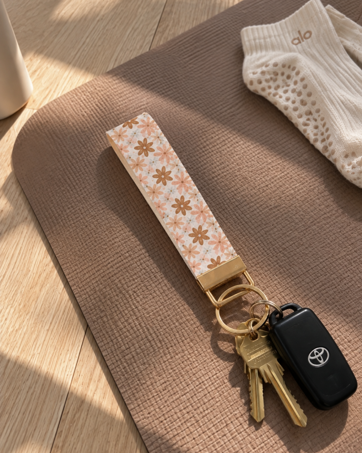 A fabric keychain with a beige and white floral pattern and gold-toned metal rings, holding car keys, laid on a brown textured blanket with a white knit sock nearby.