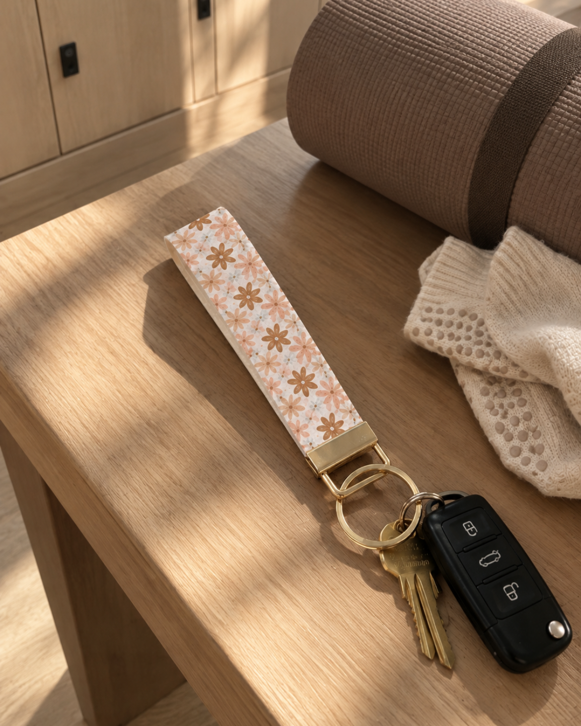 A fabric keychain strap with a beige and white floral pattern and gold metal hardware, attached to a keyring with keys and a car key fob, resting on a wooden table.