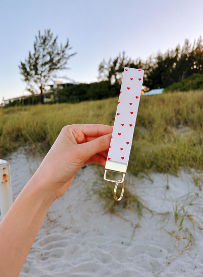 Love You Wristlet keychain with white and red heart print and gold hardware held up at the beach