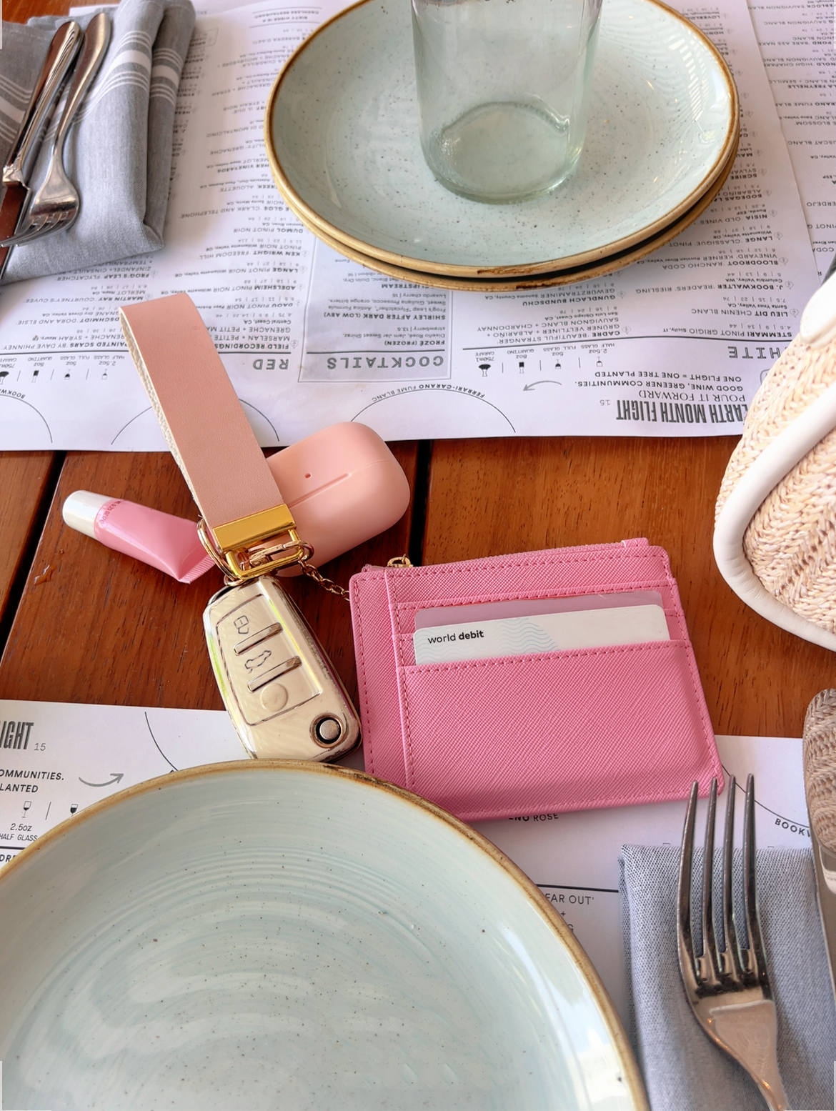 A pink textured leather card holder wallet with gold-tone hardware, shown on a wooden table surrounded by plates, a glass, a knife, and a fork.