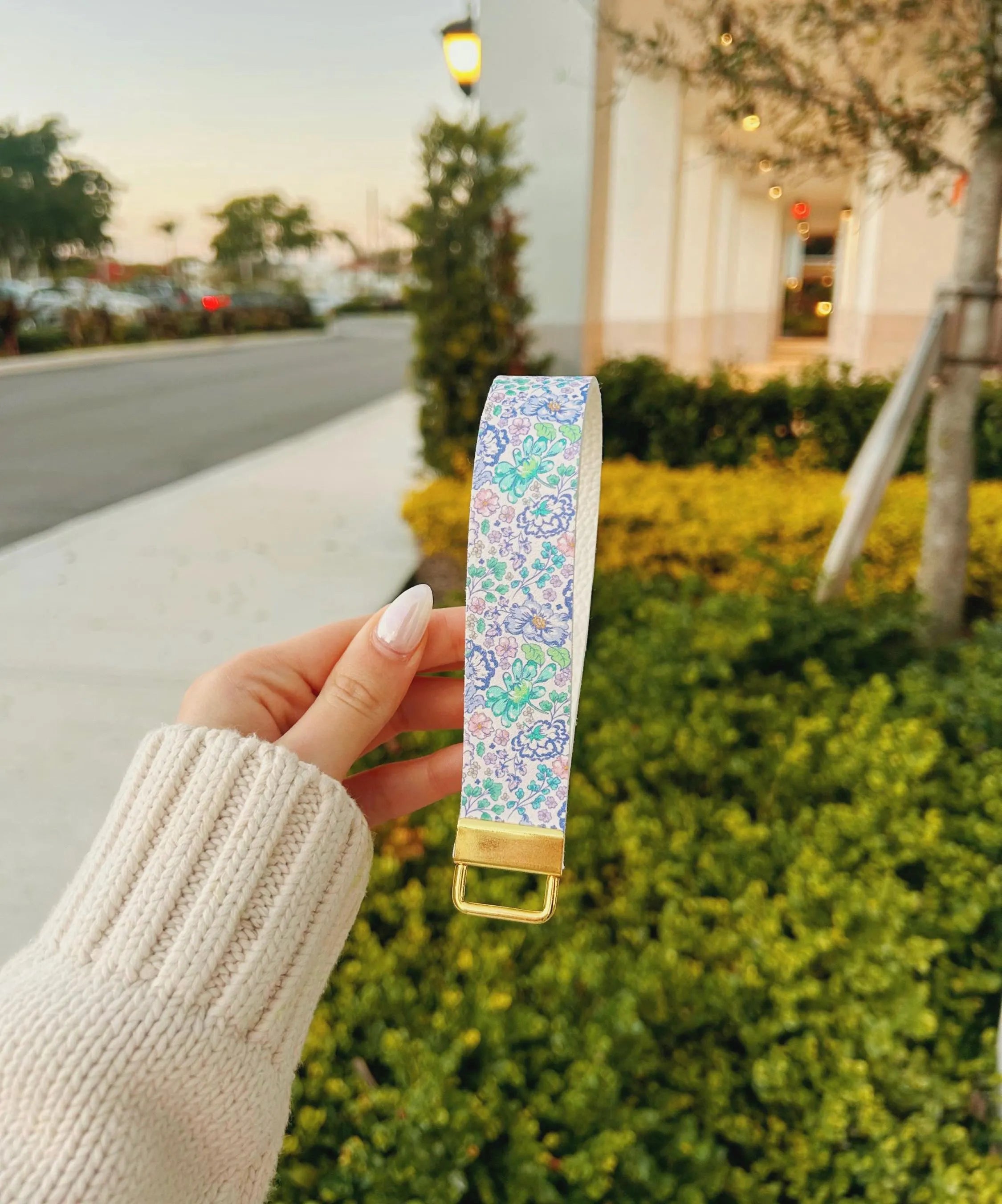 Hand holding Lavender Haze wristlet keychain with watercolor floral print in lavender, periwinkle, and mint green, outdoors at dusk with gold hardware