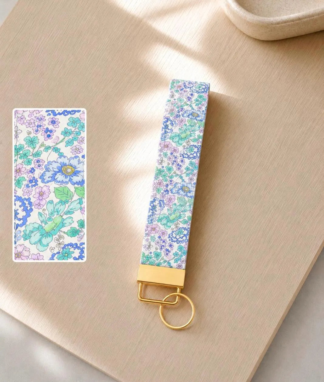 Lavender Haze wristlet keychain with watercolor floral print in lavender, mint, and periwinkle on white, laid on wood surface with gold hardware and print detail inset