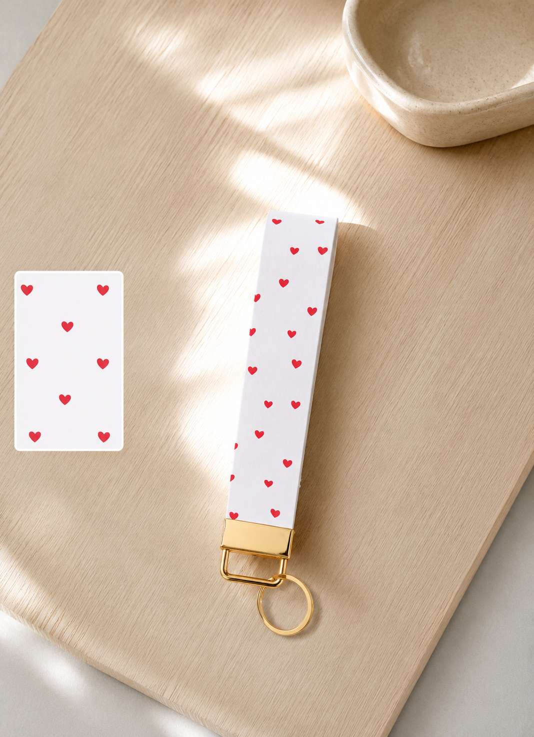 Love You Wristlet keychain with white and red heart print and gold hardware styled on a wood surface with print detail swatch