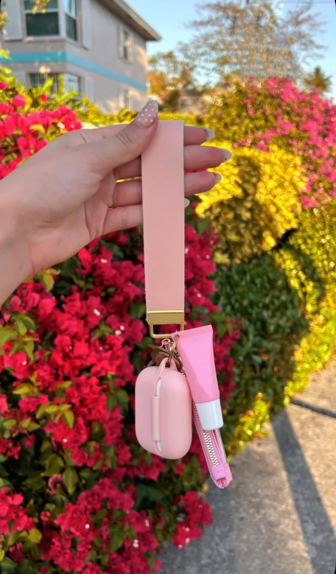 Pink wristlet wallet set with AirPods case and lip gloss held against floral background — Petal Pink Wristlet Set by Sundazed
