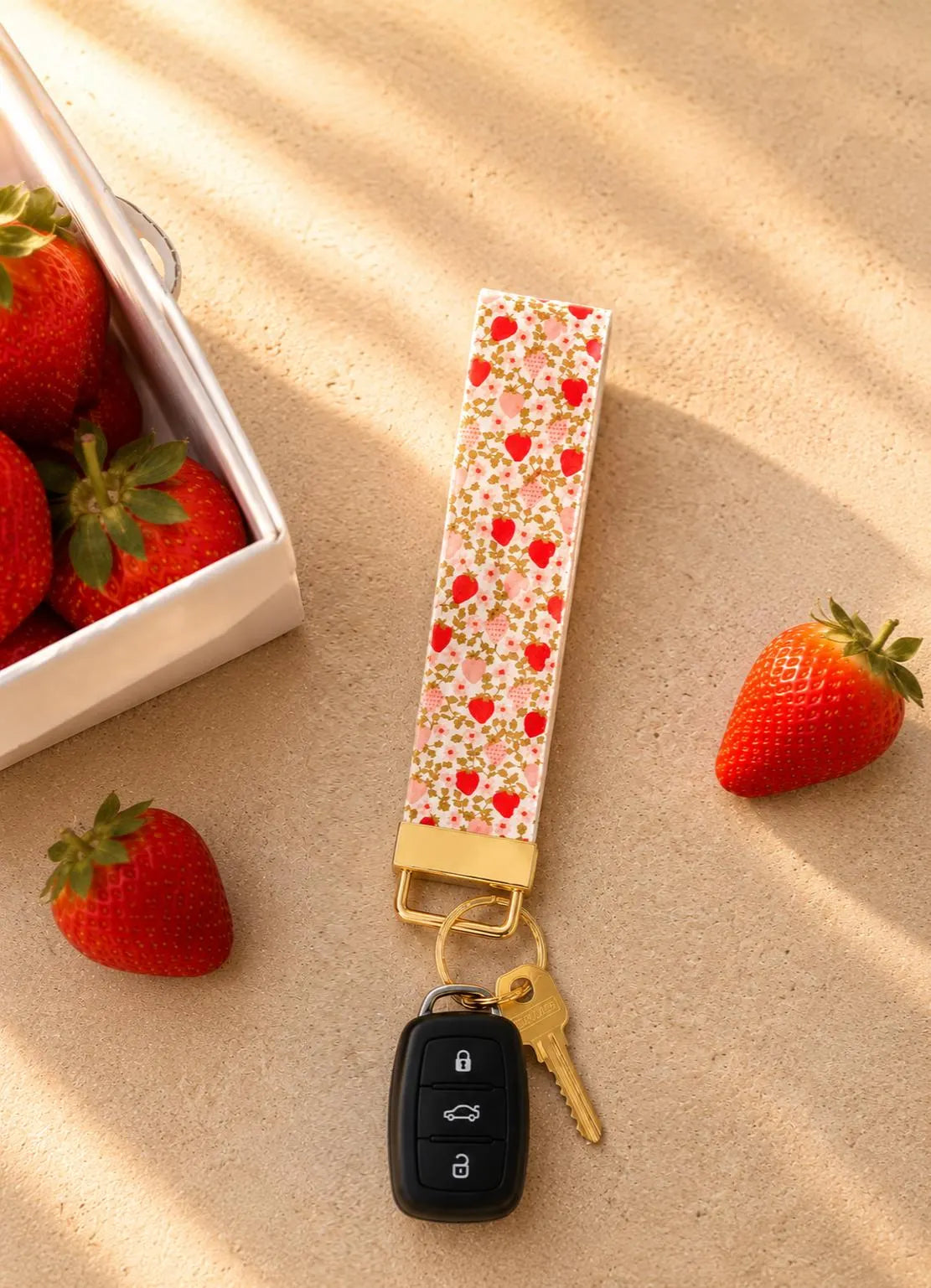 Strawberry Fields wristlet keychain with red and blush pink strawberry print on cream base, laid flat with keys attached and fresh strawberries in background, gold hardware