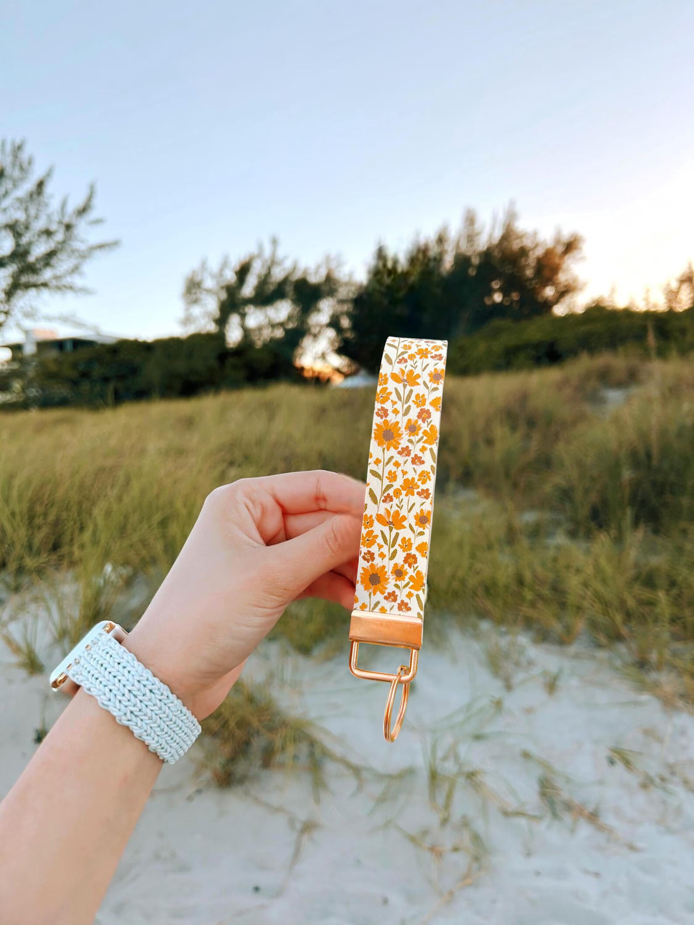 Summer Sunflower Wristlet keychain with sunflower and wildflower print on sage background with gold hardware laid flat on a light blue surface with natural light