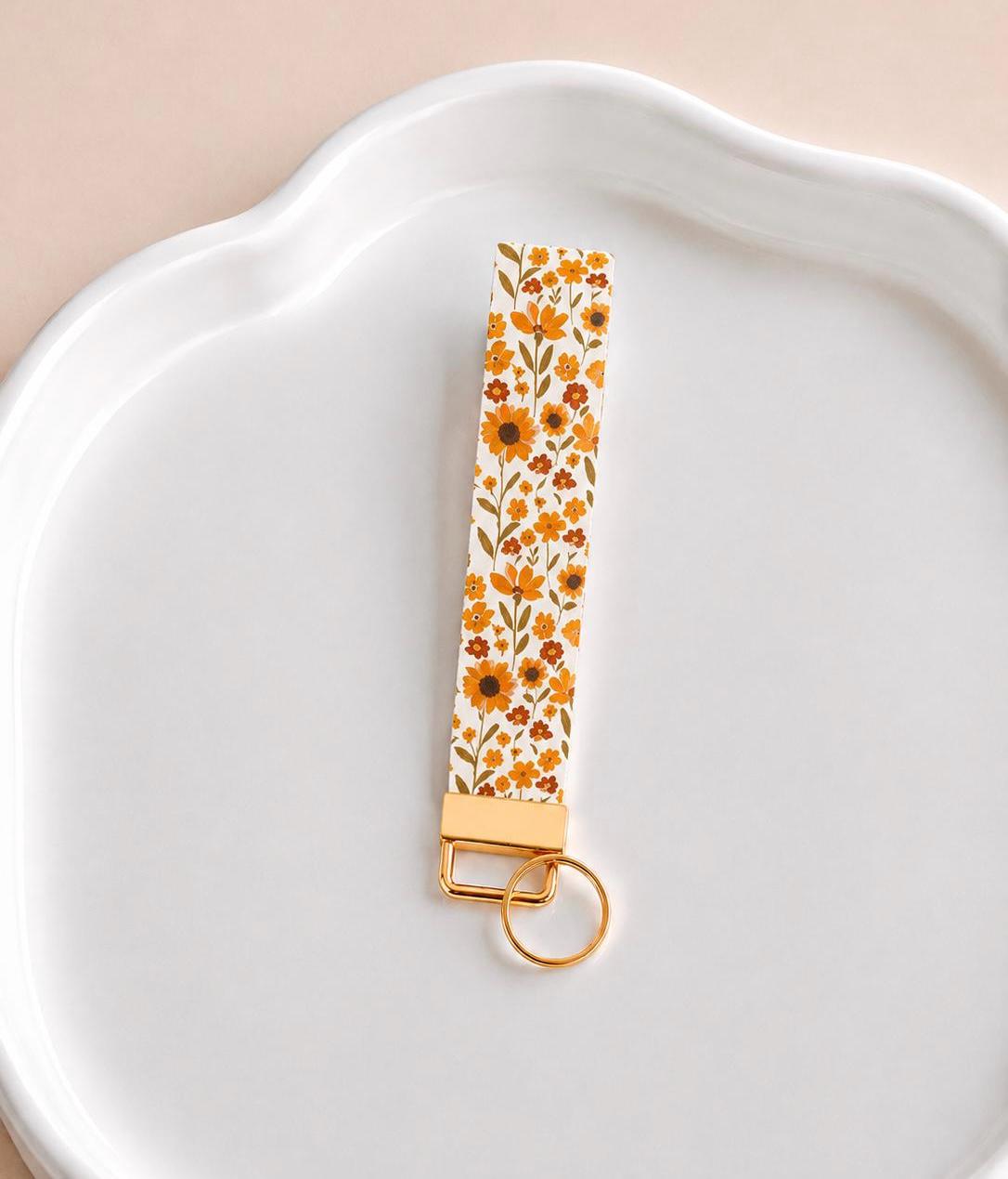 Summer Sunflower Wristlet keychain with sunflower and wildflower print on white background with gold hardware laid flat on a white ceramic surface