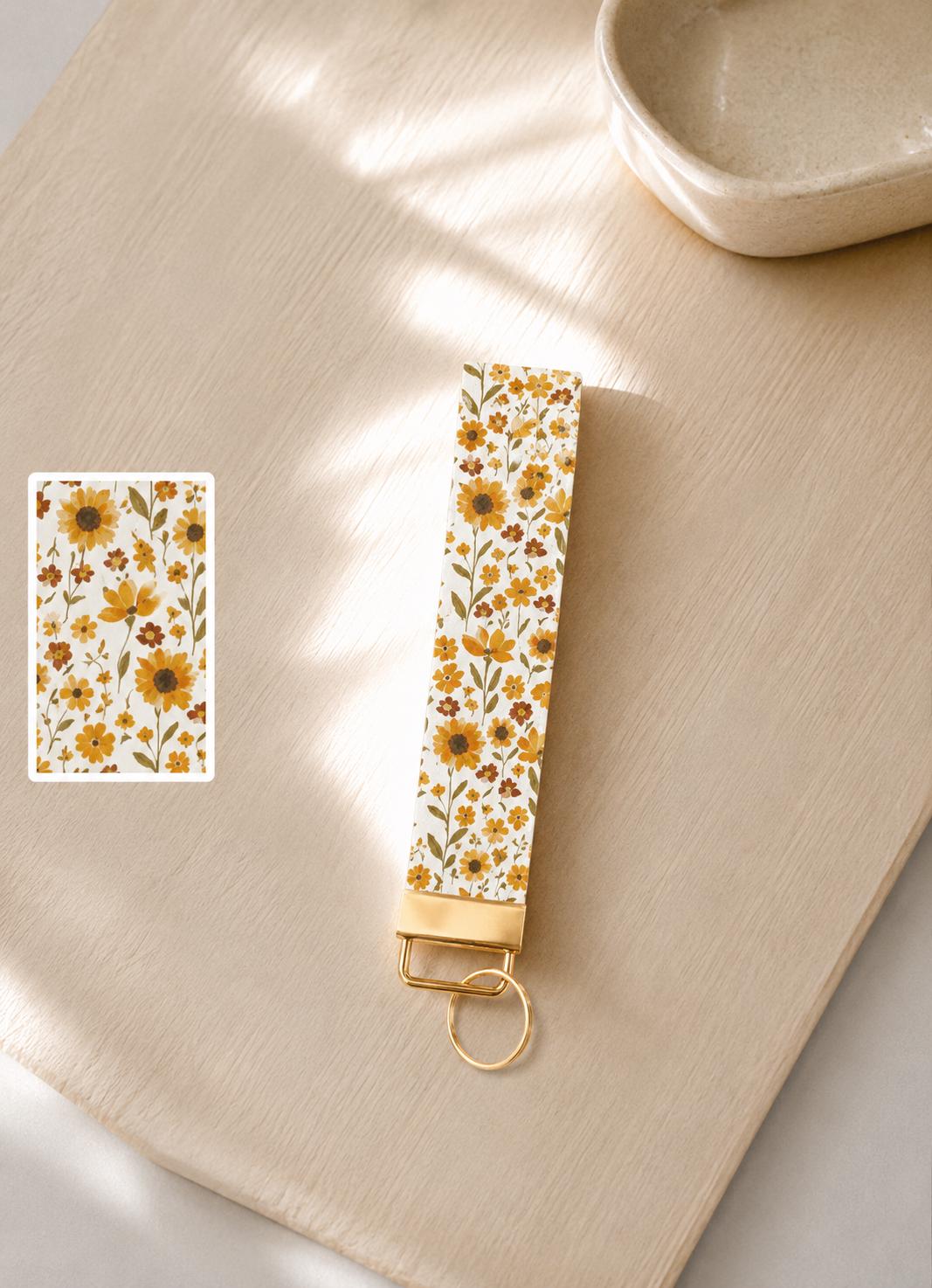 Summer Sunflower Wristlet keychain with sunflower and wildflower print on wooden light background with gold hardware laid flat on a cream surface