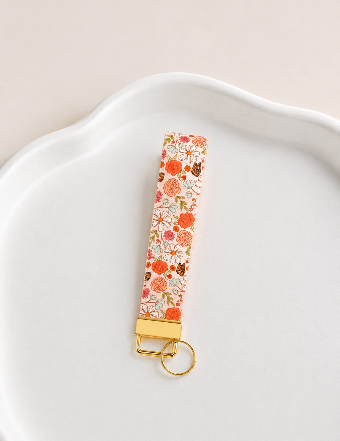 Sunkissed Skies Wristlet keychain with orange rose, daisy, and butterfly print on cream background with gold hardware on a white ceramic dish