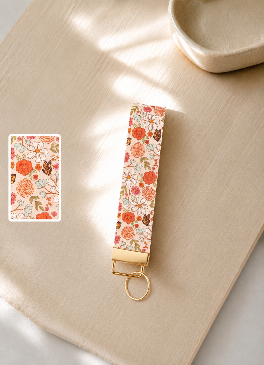 Sunkissed Skies Wristlet keychain with orange rose, daisy, and butterfly print on cream background with gold hardware styled on a wood surface with print detail swatch