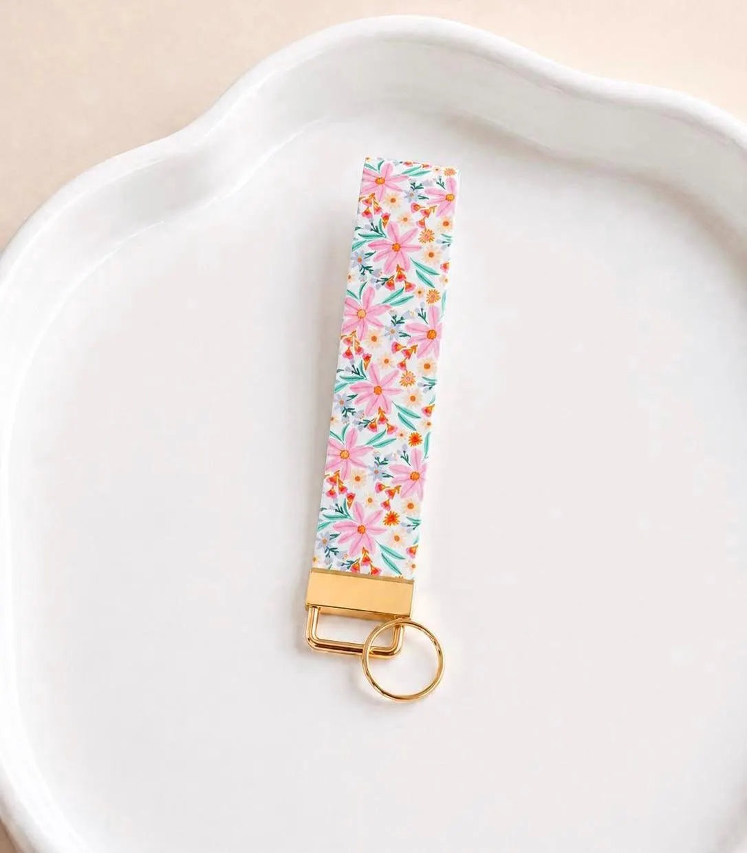 Vida Wristlet keychain with pink daisy floral print and gold hardware laid flat on a white ceramic dish