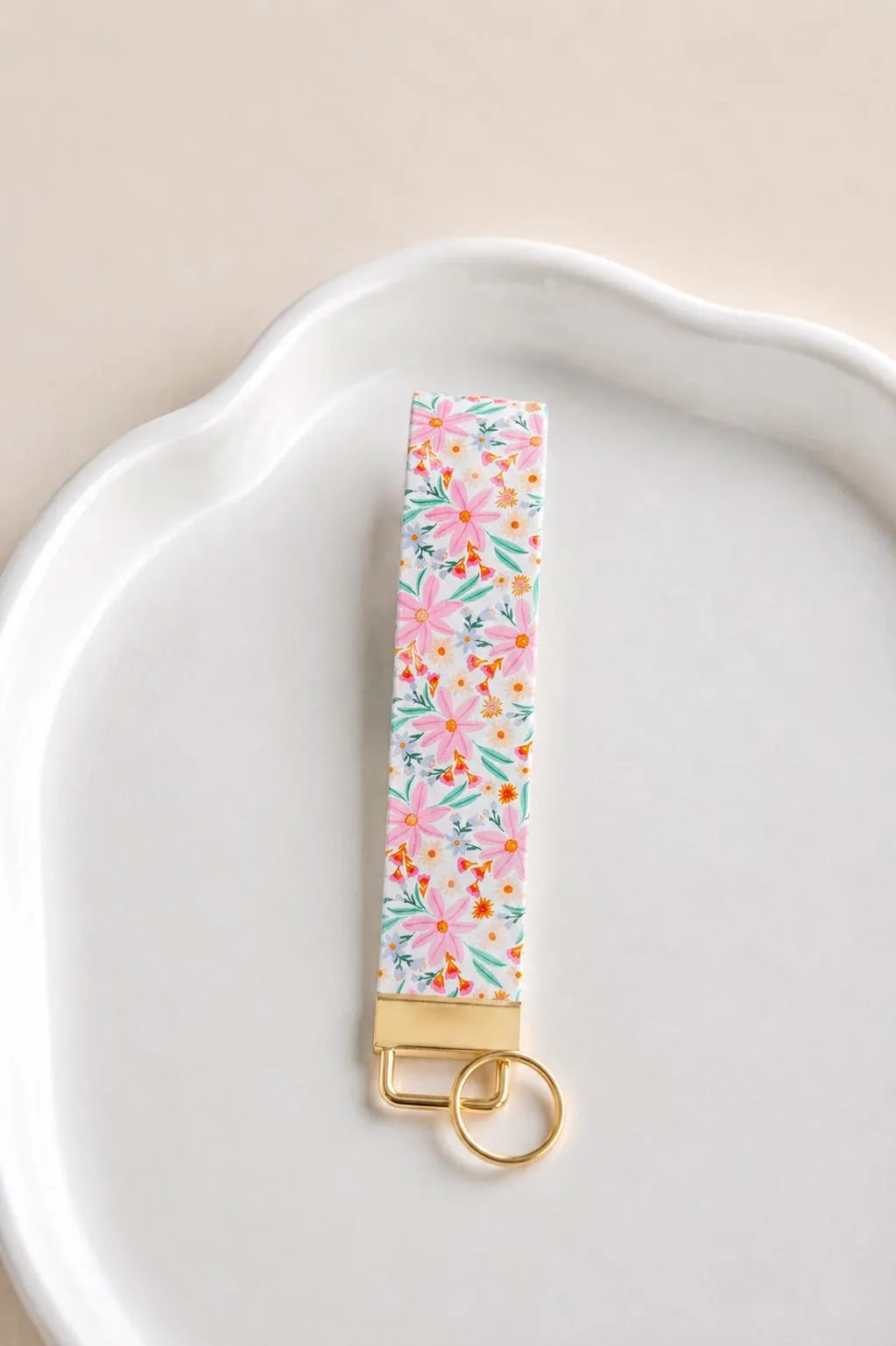 Vida Wristlet keychain with pink daisy floral print and gold hardware displayed on a white ceramic dish