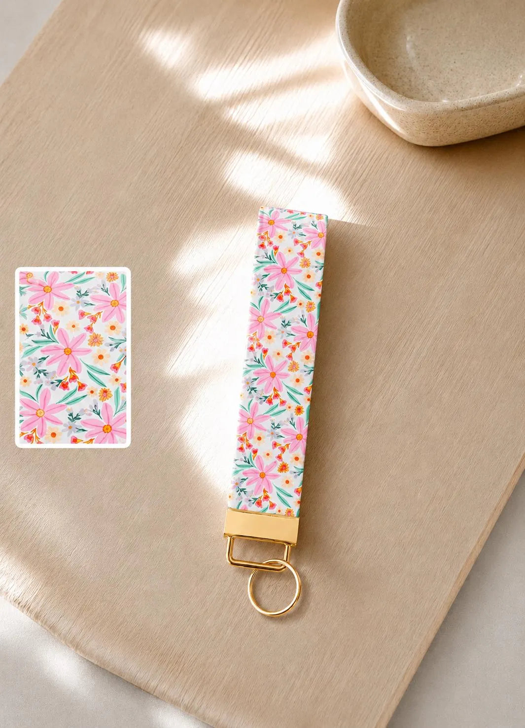 Vida Wristlet keychain with pink daisy floral print and gold hardware styled on a wooden surface.