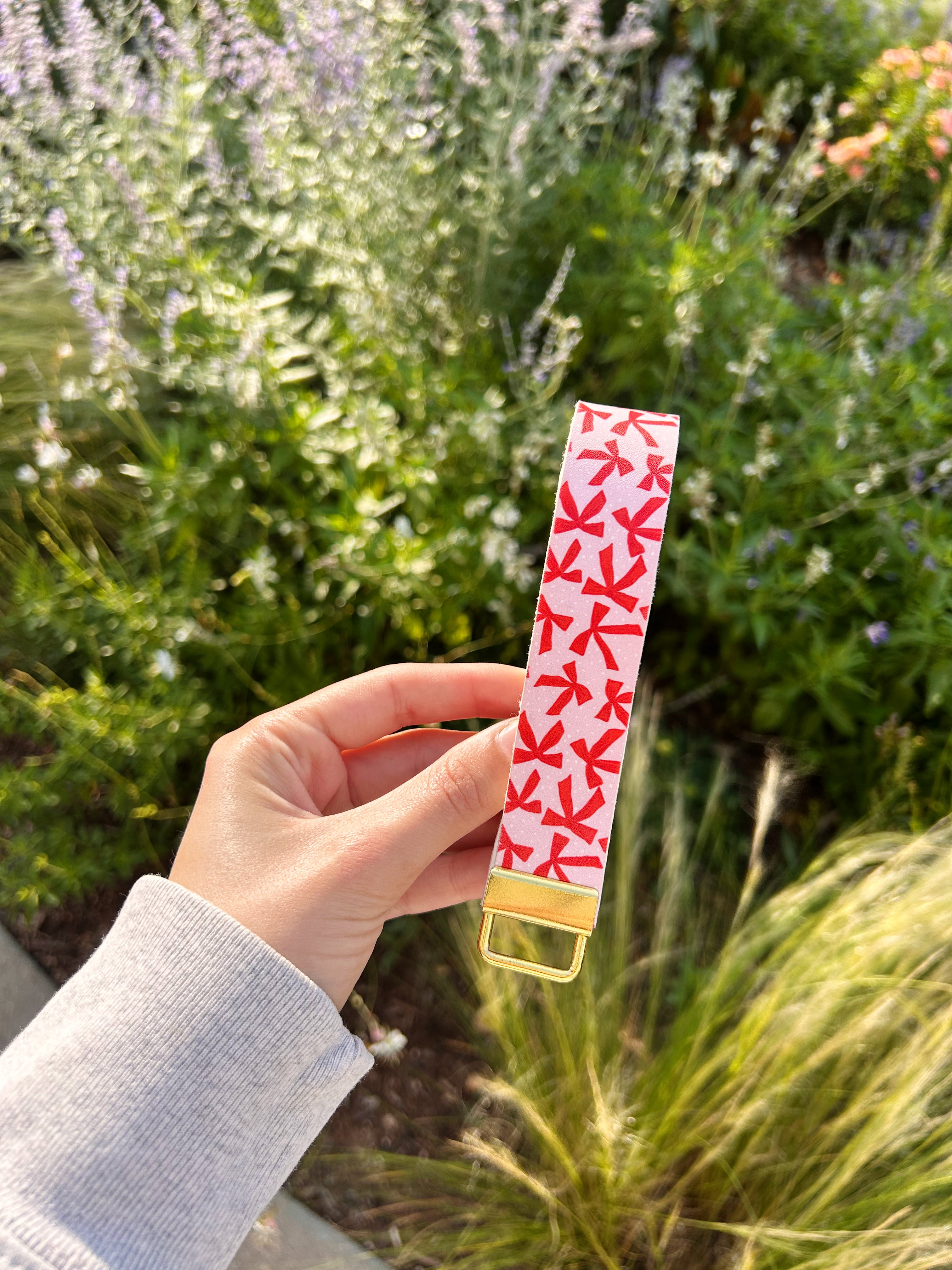 Hand holding a red and white bow patterned blush pink leather wristlet keychain for women by Sundazed — held against a garden floral background.