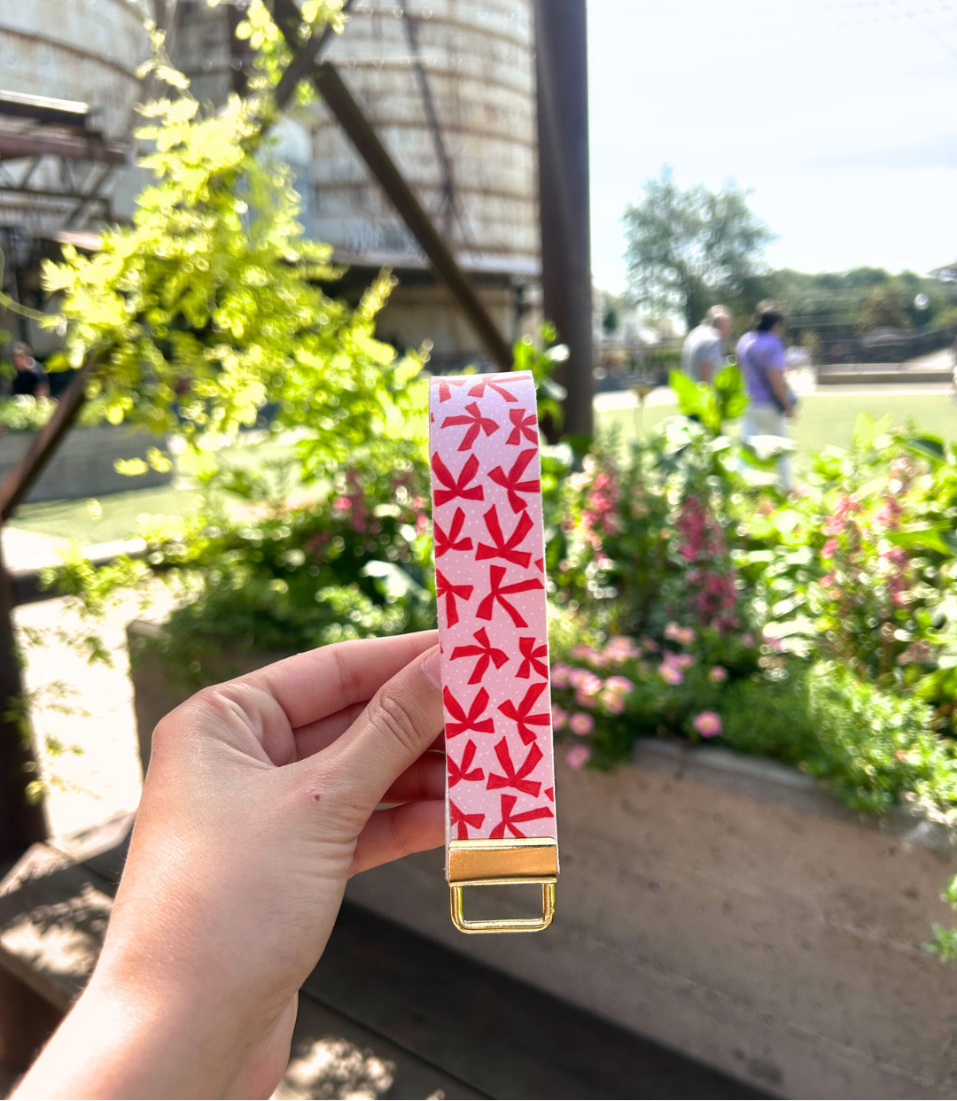 Blush pink bow-print faux leather wristlet keychain for women by Sundazed — held against a garden floral background, Bow & Go wristlet keychain.