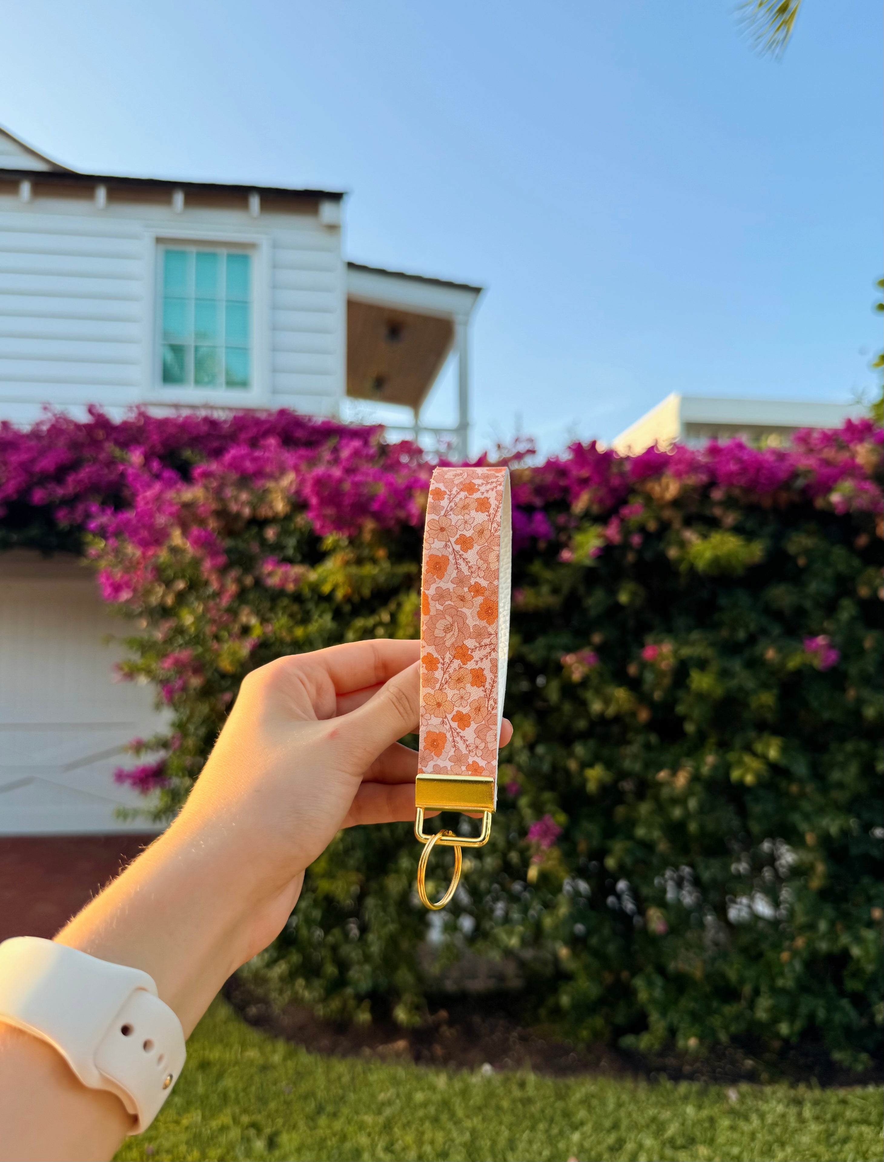 Hand holding a pink and orange patterned keychain wristlet with a gold hardware against a house and purple flowers background