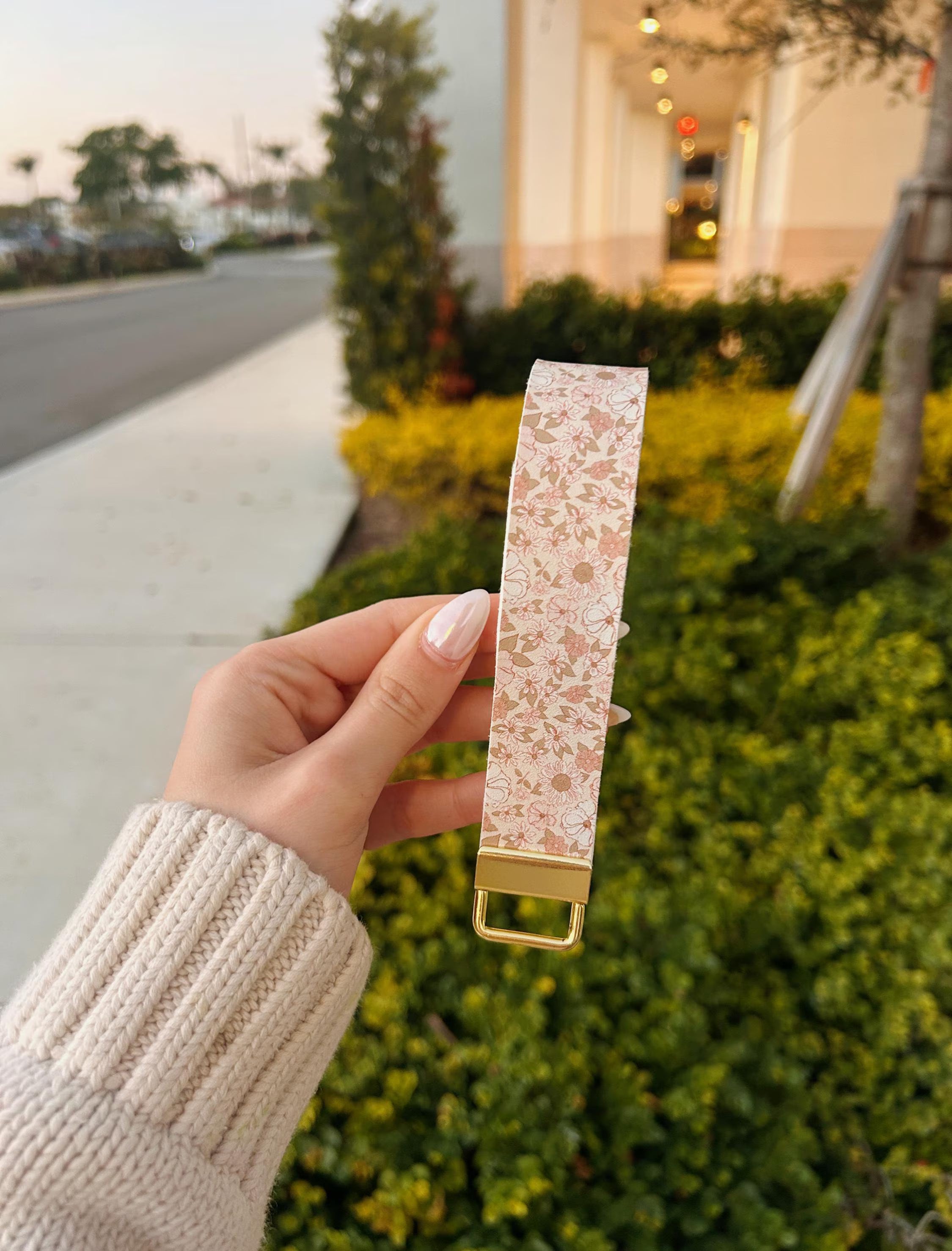 Hand holding a floral-patterned keychain wristlet in light nude pink. Standing in front of a blurred outdoor background