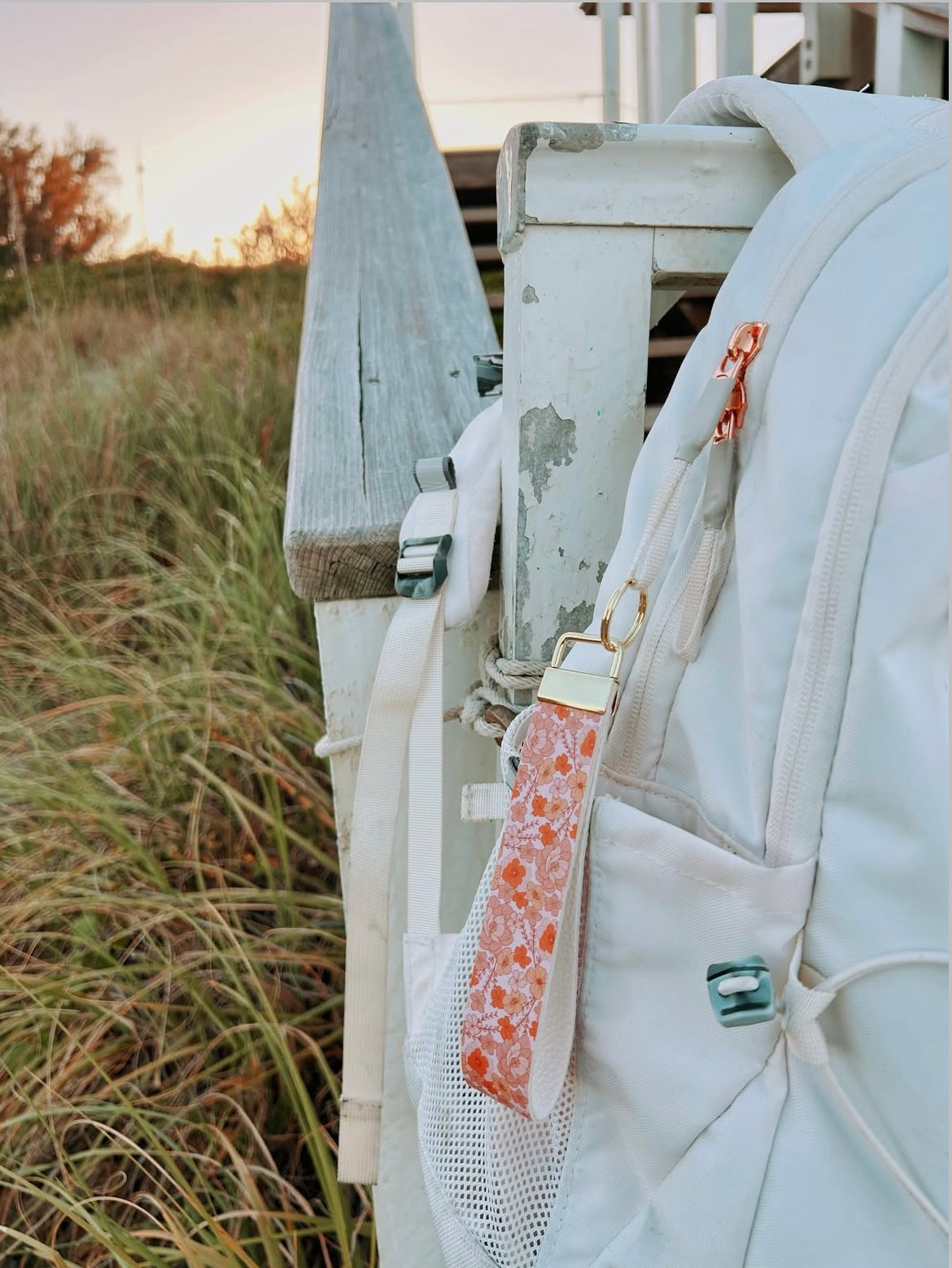 White backpack with a floral pink wristlet keychain on a wooden deck with grass in the background