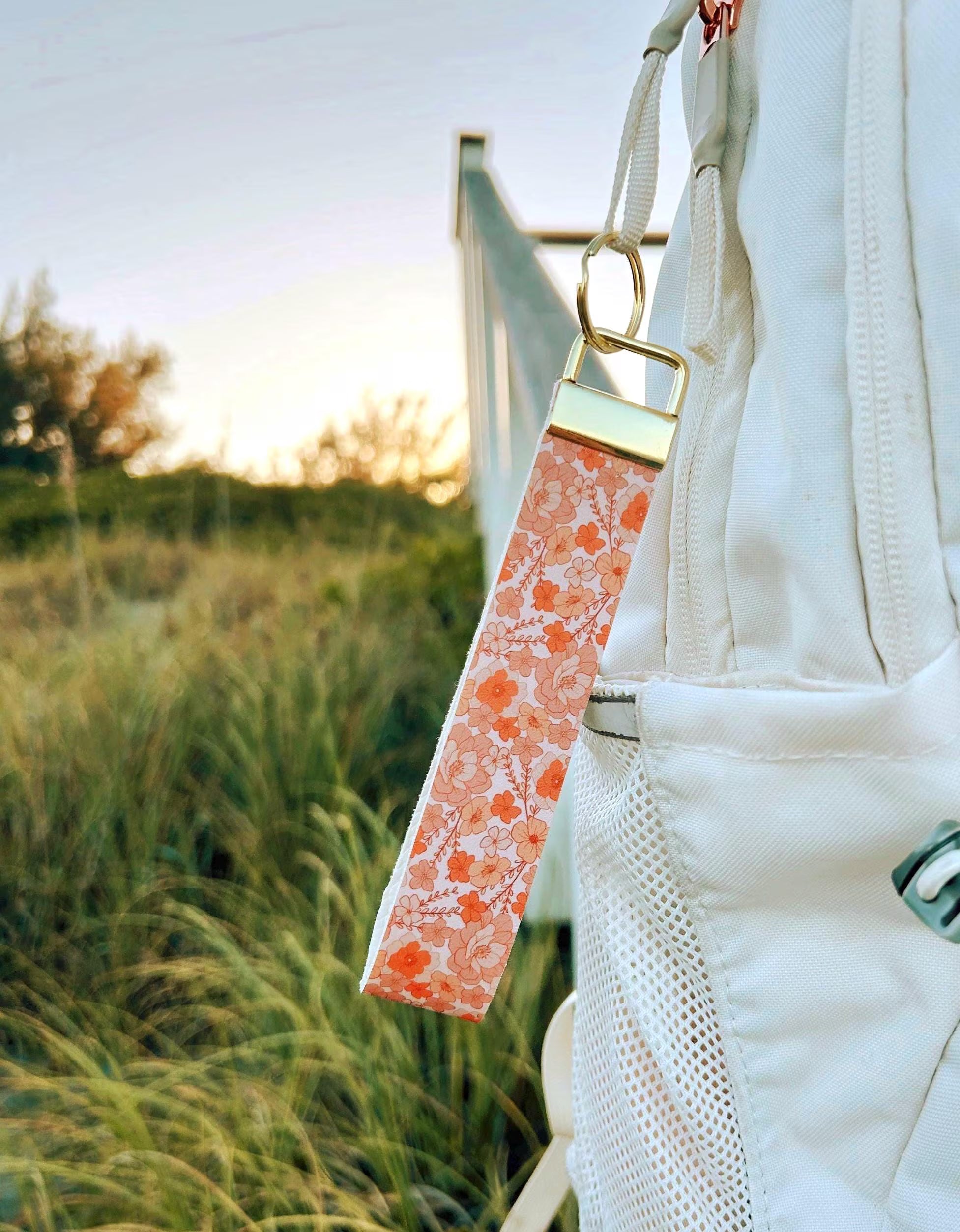 Falling for You wristlet keychain with coral and peach bloom print clipped to white bag strap outdoors at golden hour with tall grass in background