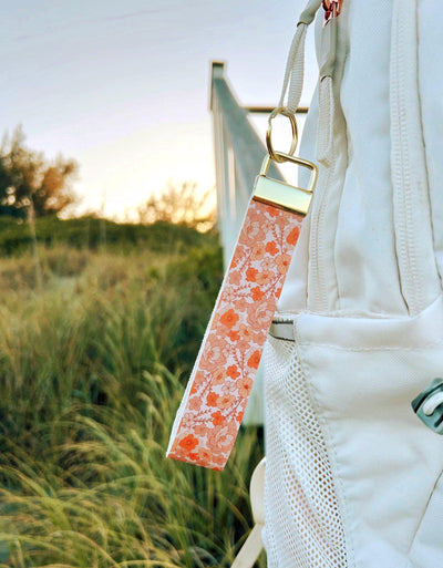 Falling for You wristlet keychain with coral and peach bloom print clipped to white bag strap outdoors at golden hour with tall grass in background
