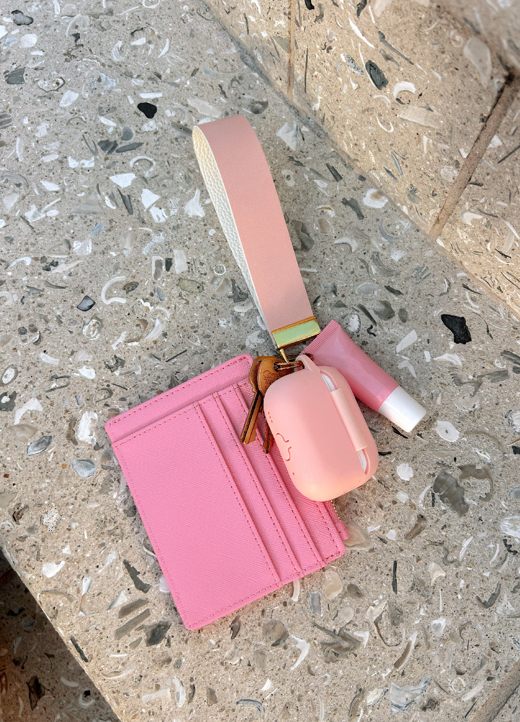 Petal pink wristlet set flat lay with cardholder wallet, AirPods case, and lip gloss on terrazzo surface — Sundazed