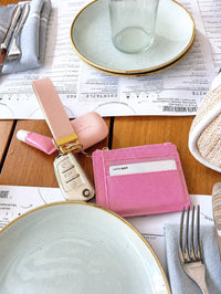 A pink leather card holder wallet with card slots and a white nameplate, displayed on a wooden table alongside a pink key fob, a car key fob, and other dining props.