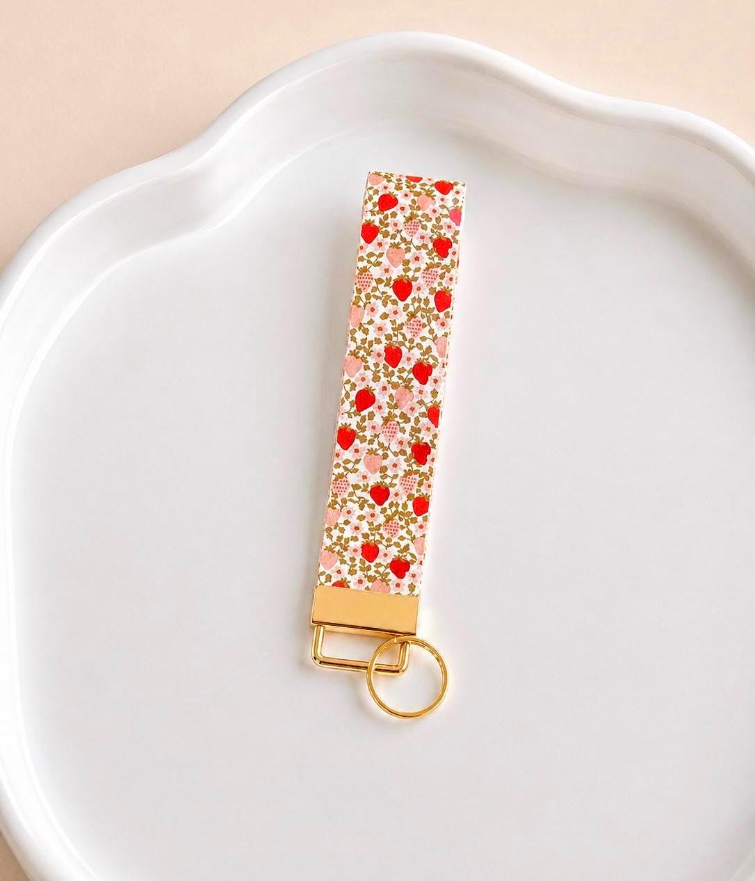 Strawberry Fields wristlet keychain with red and pink strawberry floral print on cream, laid on white scalloped tray with gold hardware