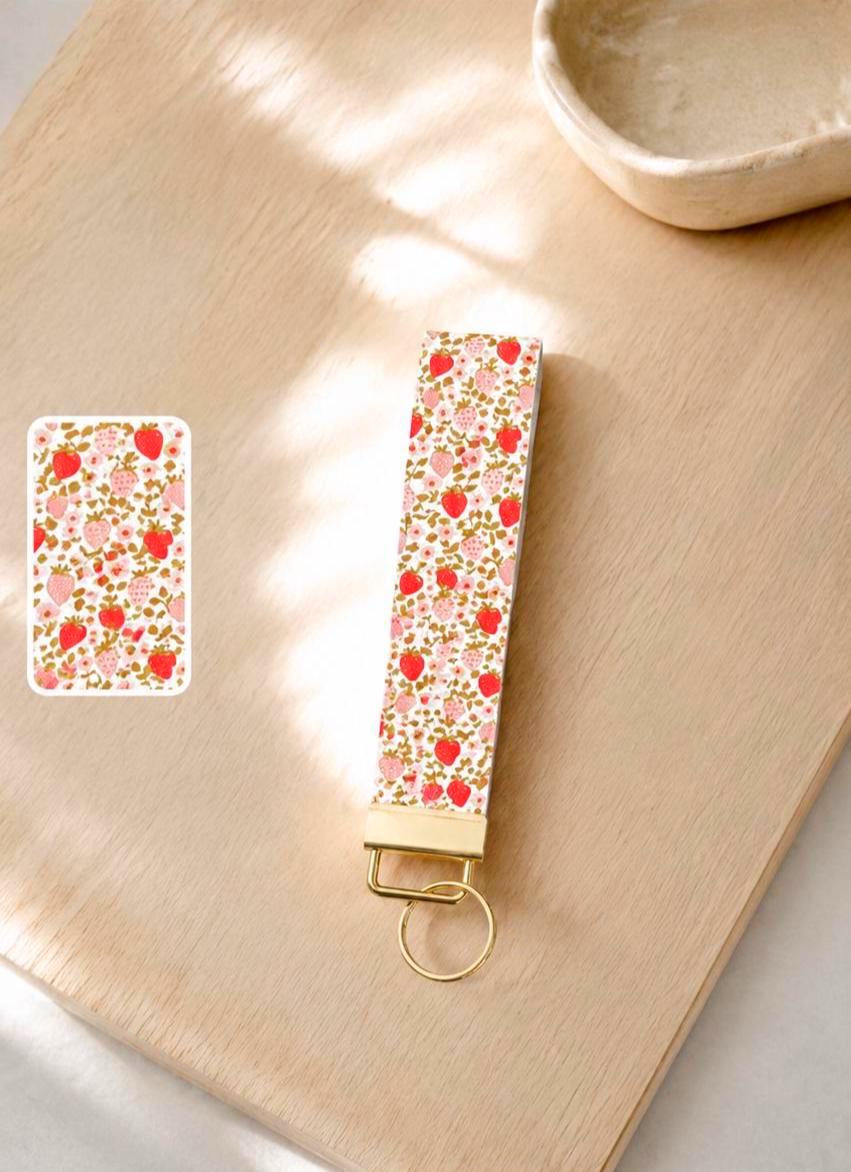 Strawberry Fields wristlet keychain with strawberry and pink floral print on cream base, laid on wood surface in natural light with print detail inset and gold hardware