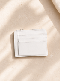 A slim white leather card holder wallet with a zip-around top and multiple card slots, photographed on a warm beige textured surface with soft shadow overlays.