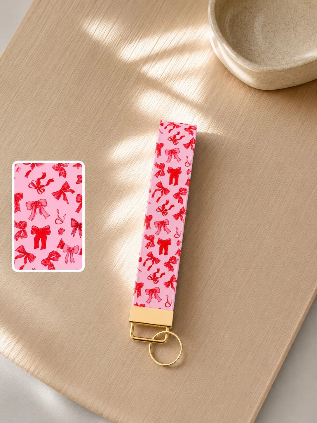 Blush Bows wristlet keychain with blush pink bow print laid on wood surface in natural light with print detail inset and gold hardware